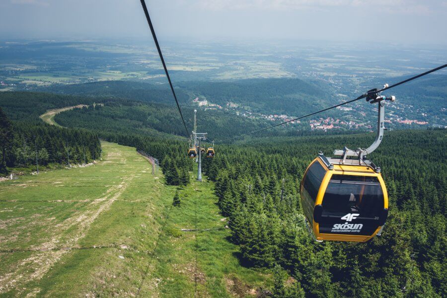 Zdjęcie. Kolej gondolowa. Na pierwszym planie wagon kolejki gondolowej. W tle inne wagony. Niżej zielona trawa oraz las. W tle panorama Świeradowa-Zdroju oraz okolicy. 