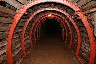 Zdjęcie. Korytarz w kopalni Liczyrzepa w Kowarach. Początkowo korytarz oświetlony. Na końcu brak oświetlenia - ciemność. Ściany korytarzu drewniane. Kilka czerwonych obręczy ciągnące się przez długość korytarza. 